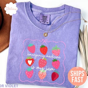 Può includere: T-shirt Comfort Colors viola con un design a fragole e la scritta "emergency medicine is my jam". La maglietta ha girocollo e maniche corte. Una collana d'oro è drappeggiata sulla maglietta.