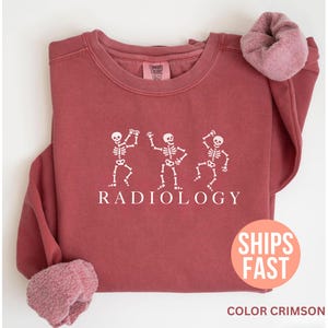 May include: Crimson-colored sweatshirt with white graphic of three dancing skeletons and the word "RADIOLOGY" below. The sweatshirt has a crew neck and long sleeves with a soft, fuzzy cuff. The image also includes a "SHIPS FAST" sticker.