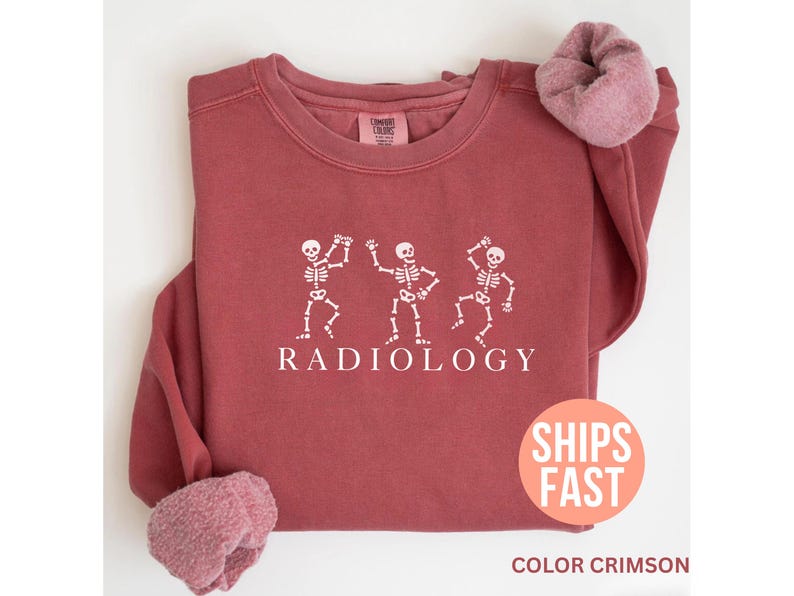 May include: Crimson-colored sweatshirt with white graphic of three dancing skeletons and the word "RADIOLOGY" below. The sweatshirt has a crew neck and long sleeves with a soft, fuzzy cuff. The image also includes a "SHIPS FAST" sticker.