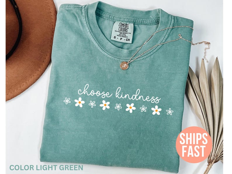 May include: Light green t-shirt with the phrase "choose kindness" in white script and daisy graphics. A silver necklace with a pendant rests on the shirt. The shirt is folded and has a Comfort Colors tag.