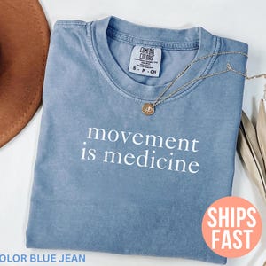 K&ouml;nnte beinhalten: Blaues Jeans-T-Shirt mit dem wei&szlig;en Schriftzug &bdquo;movement is medicine&ldquo;. Eine goldene Halskette mit Anh&auml;nger liegt auf dem Shirt. Das Shirt ist auf einer wei&szlig;en Oberfl&auml;che gefaltet.