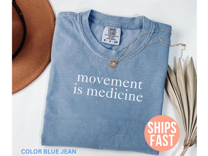 K&ouml;nnte beinhalten: Blaues Jeans-T-Shirt mit dem wei&szlig;en Schriftzug &bdquo;movement is medicine&ldquo;. Eine goldene Halskette mit Anh&auml;nger liegt auf dem Shirt. Das Shirt ist auf einer wei&szlig;en Oberfl&auml;che gefaltet.