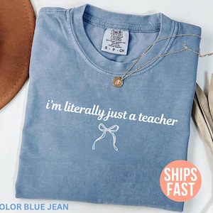 May include: Blue jean-colored t-shirt with the text "i'm literally just a teacher" in white. A gold necklace with a pendant is draped across the shirt. A brown hat and dried leaves are in the background.