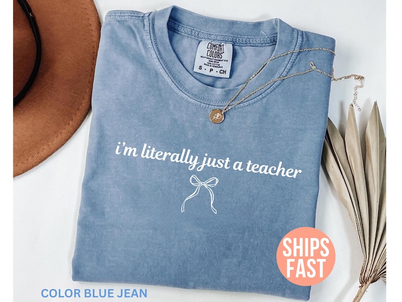 May include: Blue jean-colored t-shirt with the text "i'm literally just a teacher" in white. A gold necklace with a pendant is draped across the shirt. A brown hat and dried leaves are in the background.