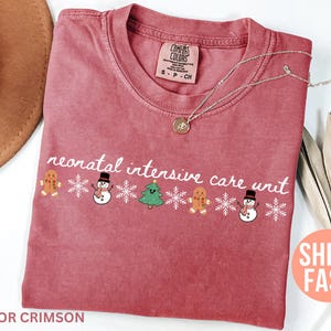 May include: Crimson-colored t-shirt with the text "neonatal intensive care unit" in white script. The design includes gingerbread men, snowmen, snowflakes, and a Christmas tree. A silver necklace with a pendant is on the shirt.