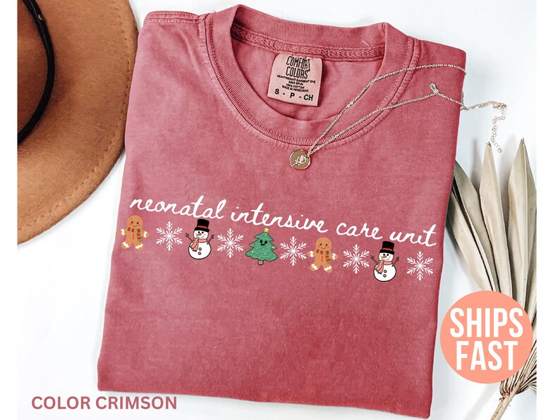 May include: Crimson-colored t-shirt with the text "neonatal intensive care unit" in white script. The design includes gingerbread men, snowmen, snowflakes, and a Christmas tree. A silver necklace with a pendant is on the shirt.
