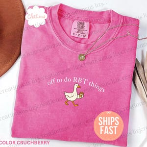 May include: A bright pink short-sleeve t-shirt featuring a graphic of a white goose holding a coffee cup. The text above the goose reads off to do RBT things. The shirt has a crew neckline and a Comfort Colors brand label inside the collar.