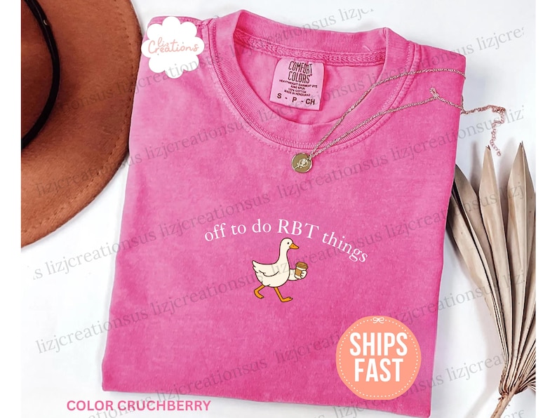 May include: A bright pink short-sleeve t-shirt featuring a graphic of a white goose holding a coffee cup. The text above the goose reads off to do RBT things. The shirt has a crew neckline and a Comfort Colors brand label inside the collar.