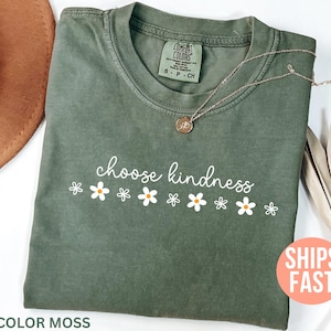 May include: A moss green t-shirt with the words "choose kindness" in white script, accompanied by white daisy graphics. A gold necklace with a pendant rests on the shirt. The shirt is labeled "Comfort Colors".