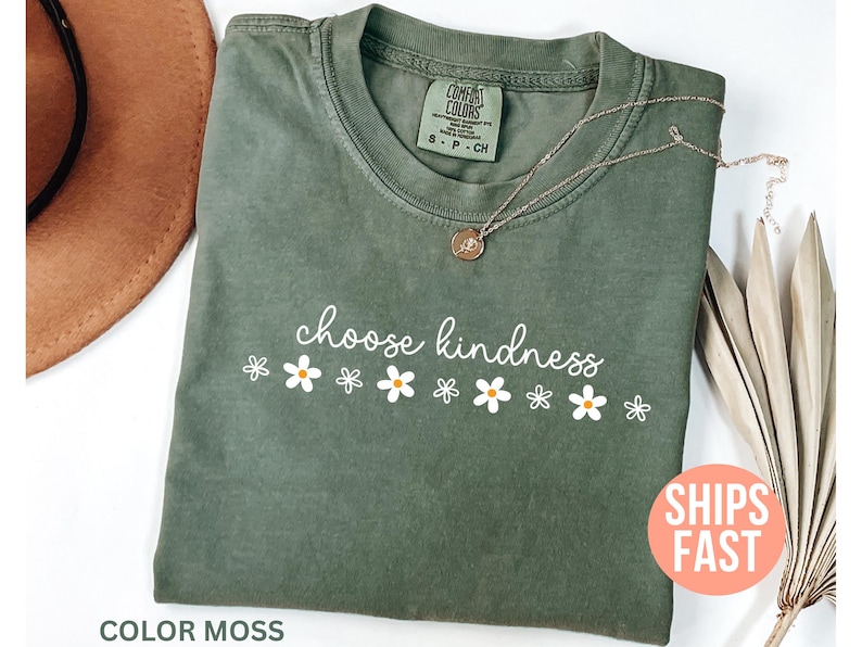 May include: A moss green t-shirt with the words "choose kindness" in white script, accompanied by white daisy graphics. A gold necklace with a pendant rests on the shirt. The shirt is labeled "Comfort Colors".