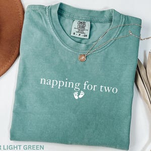 Puede incluir: Camiseta verde claro con el texto "napping for two" y huellas de beb&eacute;. Un collar plateado con un colgante descansa sobre la camiseta. Un sombrero marr&oacute;n y una planta seca est&aacute;n en el fondo.