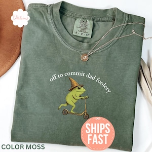 Puede incluir: Una camiseta de color musgo con la frase "off to commit dad foolery" y una rana de dibujos animados montada en un scooter, con un sombrero de mago. Un collar de oro descansa sobre la camiseta. La camiseta tiene cuello redondo y mangas cortas.