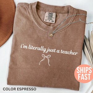 May include: Espresso-colored t-shirt with the text "i'm literally just a teacher" in white. A silver necklace with a pendant rests on the shirt. The shirt has a crew neck and a small tag.