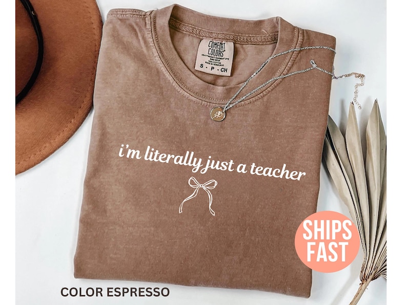 May include: Espresso-colored t-shirt with the text "i'm literally just a teacher" in white. A silver necklace with a pendant rests on the shirt. The shirt has a crew neck and a small tag.