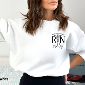 May include: White sweatshirt with a black graphic of a medical caduceus and the letters "RN" with the name "Ashley" underneath.