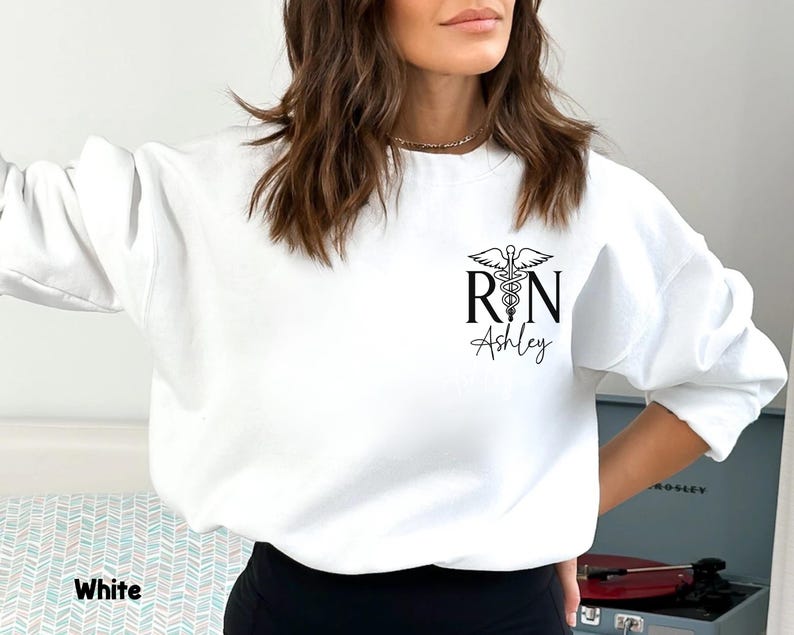 May include: White sweatshirt with a black graphic of a medical caduceus and the letters "RN" with the name "Ashley" underneath.