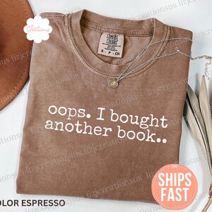 May include: An espresso-colored short-sleeve t-shirt with a crew neckline. The shirt features the white text "oops. I bought another book.." printed across the chest. A silver-toned necklace with a small circular pendant rests on the collar.