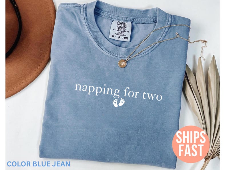 Puede incluir: Una camiseta azul vaquero con el texto "napping for two" y huellas de beb&eacute;. Un collar de oro con un colgante est&aacute; en la camisa. La camisa est&aacute; doblada sobre una superficie blanca con un sombrero marr&oacute;n y una planta seca.