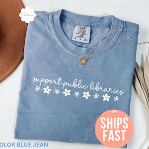 May include: A blue jean colored t-shirt with the text "support public libraries" in white script, decorated with small daisy graphics. A gold necklace and a brown hat are visible. The shirt has a "Ships Fast" sticker.