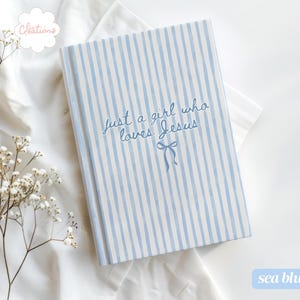 May include: A sea blue notebook with vertical white stripes. The cover features the text "Just a girl who loves Jesus" in cursive script, with a small bow illustration. The notebook is on a white surface with floral accents.