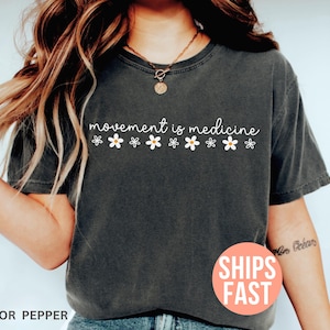 May include: Dark gray t-shirt with the text "movement is medicine" in white script, decorated with small daisy graphics. The shirt is a solid color and has a relaxed fit. The color is described as "pepper".