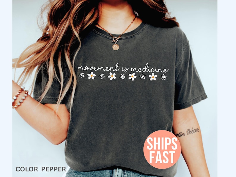 May include: Dark gray t-shirt with the text "movement is medicine" in white script, decorated with small daisy graphics. The shirt is a solid color and has a relaxed fit. The color is described as "pepper".
