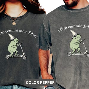 Puede incluir: Dos camisetas de color gris oscuro con gráficos a juego. Cada camiseta presenta una rana verde claro con un sombrero de mago y montada en un patinete, con el texto "off to commit mom folery" y "off to commit dad folery" encima de la rana.
