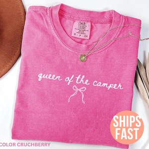 May include: A bright pink t-shirt with the words "queen of the camper" in white script. A gold necklace with a pendant rests on the shirt. The shirt has a crew neck and short sleeves. The shirt is labeled "Comfort Colors".