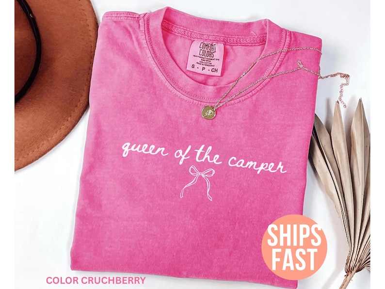 May include: A bright pink t-shirt with the words "queen of the camper" in white script. A gold necklace with a pendant rests on the shirt. The shirt has a crew neck and short sleeves. The shirt is labeled "Comfort Colors".