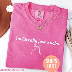 Pode incluir: Uma t-shirt rosa brilhante com o texto branco "i'm literally just a bcba" e um laço. Um colar dourado e um chapéu castanho também são visíveis. A t-shirt está rotulada como "Color Crunchberry".