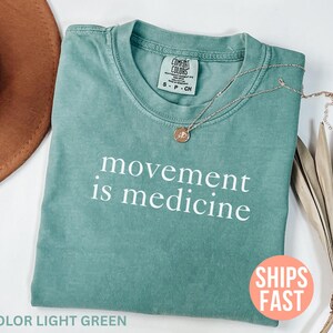 K&ouml;nnte beinhalten: Ein hellgr&uuml;nes T-Shirt mit dem wei&szlig;en Aufdruck &bdquo;movement is medicine&ldquo;. Eine goldene Halskette mit Anh&auml;nger liegt auf dem Shirt. Ein brauner Hut und eine getrocknete Pflanze befinden sich im Hintergrund.