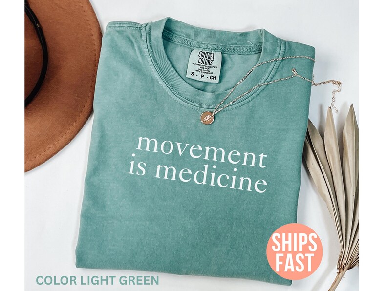 K&ouml;nnte beinhalten: Ein hellgr&uuml;nes T-Shirt mit dem wei&szlig;en Aufdruck &bdquo;movement is medicine&ldquo;. Eine goldene Halskette mit Anh&auml;nger liegt auf dem Shirt. Ein brauner Hut und eine getrocknete Pflanze befinden sich im Hintergrund.