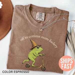 May include: Espresso-coloured t-shirt with a green frog on a scooter, wearing a wizard hat. The text "off to commit mom foolery" is above the frog. A silver necklace is draped across the shirt.