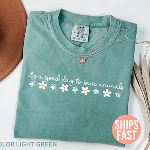 K&ouml;nnte beinhalten: Hellgr&uuml;nes T-Shirt mit dem Schriftzug "it's a good day to save animals" und G&auml;nsebl&uuml;mchen-Grafiken. Eine silberne Halskette mit Anh&auml;nger liegt auf dem Shirt. Das Shirt ist gefaltet, mit einem braunen Hut im Hintergrund.