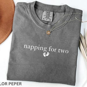 Puede incluir: Una camiseta gris Comfort Colors con el texto "napping for two" y huellas de beb&eacute;. Tambi&eacute;n se ven un collar con colgante dorado y un sombrero marr&oacute;n. La camiseta est&aacute; doblada cuidadosamente.