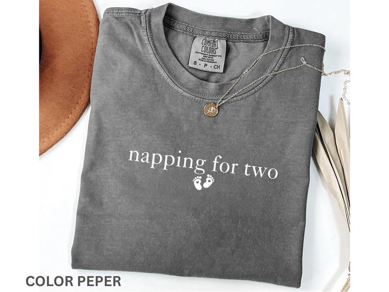 Puede incluir: Una camiseta gris Comfort Colors con el texto "napping for two" y huellas de beb&eacute;. Tambi&eacute;n se ven un collar con colgante dorado y un sombrero marr&oacute;n. La camiseta est&aacute; doblada cuidadosamente.