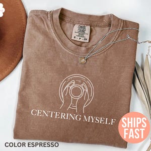 Könnte beinhalten: Espressofarbenes T-Shirt mit dem weißen Schriftzug "CENTERING MYSELF". Das Shirt zeigt eine Grafik von Händen, die eine Tasse halten. Eine silberne Halskette mit einem kleinen Anhänger liegt auf dem Shirt.