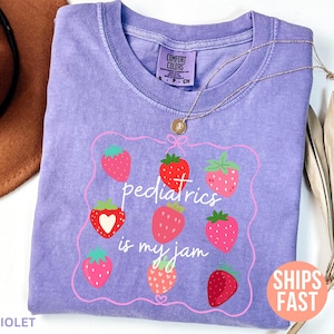 May include: A lilac Comfort Colors t-shirt featuring a strawberry design and the phrase "pediatrics is my jam." The design includes a pink frame with red and pink strawberries. A gold necklace is draped over the shirt.