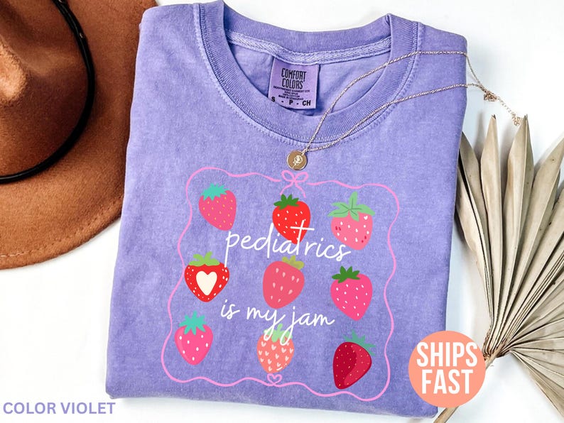 May include: A lilac Comfort Colors t-shirt featuring a strawberry design and the phrase "pediatrics is my jam." The design includes a pink frame with red and pink strawberries. A gold necklace is draped over the shirt.