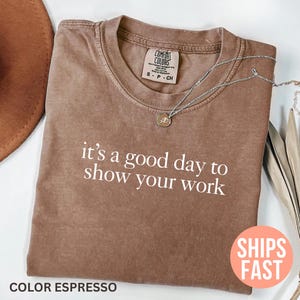 Könnte beinhalten: Espressofarbenes T-Shirt mit dem weißen Text "it's a good day to show your work". Das Shirt hat einen Rundhalsausschnitt und kurze Ärmel. Eine silberne Halskette mit Anhänger befindet sich auf dem Shirt. Ein brauner Hut ist im Hintergrund.