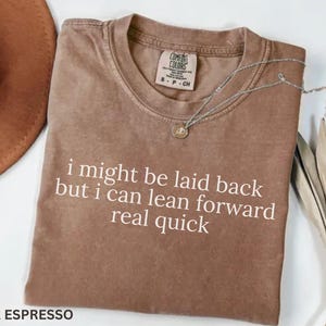 May include: Espresso-colored t-shirt with the text "i might be laid back but i can lean forward real quick" in white. A silver necklace with a pendant rests on the shirt. A brown hat and dried leaves are in the background.