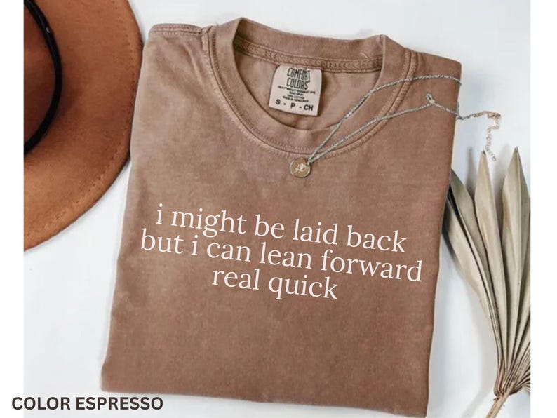 May include: Espresso-colored t-shirt with the text "i might be laid back but i can lean forward real quick" in white. A silver necklace with a pendant rests on the shirt. A brown hat and dried leaves are in the background.