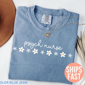 Op de afbeelding: Blauw spijkerbroekkleurig T-shirt met de woorden "psych nurse" in witte letters, versierd met witte en gele madeliefjes. Een gouden ketting met een hanger ligt op het shirt. Het shirt heeft een "Comfort Colors" label.