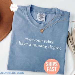 Pode incluir: Uma camiseta azul jeans com o texto "everyone relax i have a nursing degree" em branco. A camisa tem gola redonda e mangas curtas. Um colar de ouro com um pingente está sobre a camisa.