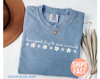 May include: Blue jean colored t-shirt with the phrase "it's a good day to save animals" in white script and daisy graphics. A gold necklace with a pendant is on the shirt. The shirt has a Comfort Colors tag.