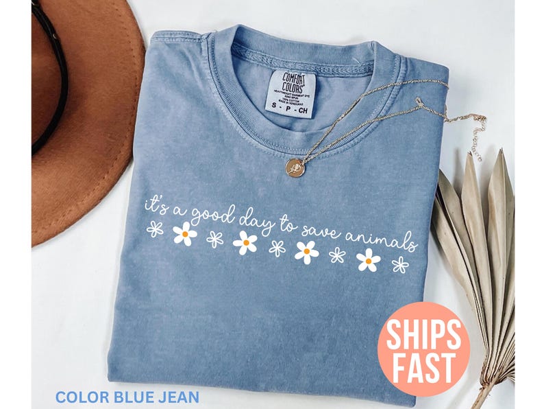 K&ouml;nnte beinhalten: Blaues Jeans-T-Shirt mit dem wei&szlig;en Schriftzug "it's a good day to save animals" und G&auml;nsebl&uuml;mchen-Grafiken. Eine goldene Halskette mit Anh&auml;nger befindet sich auf dem Shirt. Das Shirt hat ein Comfort Colors-Etikett.