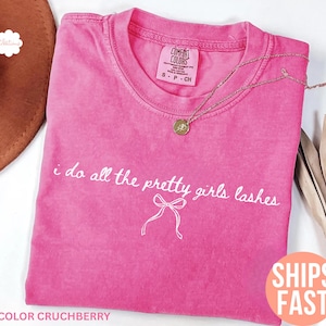 May include: A pink Comfort Colors t-shirt with the text "i do all the pretty girls lashes" in white script. A gold necklace with a pendant rests on the shirt. The shirt is folded, and a brown hat is partially visible.