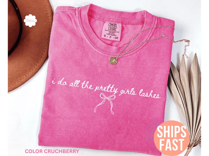 May include: A pink Comfort Colors t-shirt with the text "i do all the pretty girls lashes" in white script. A gold necklace with a pendant rests on the shirt. The shirt is folded, and a brown hat is partially visible.
