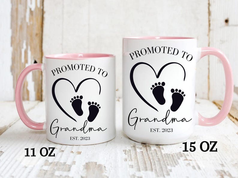K&ouml;nnte beinhalten: Zwei wei&szlig;e Keramikbecher mit rosa R&auml;ndern. Die Becher haben schwarzen Text, der "Bef&ouml;rdert zur Oma Est. 2023" lautet, mit einem schwarzen Herzen und einem Fu&szlig;abdruckdesign. Der Becher links ist 11 oz und der Becher rechts ist 15 oz.