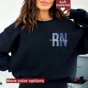 May include: Black sweatshirt with the letters "RN" in blue and the name "Ashley" in a script font. The sweatshirt has a crew neck and long sleeves. The words "Soft material" and "More color options" are also visible.